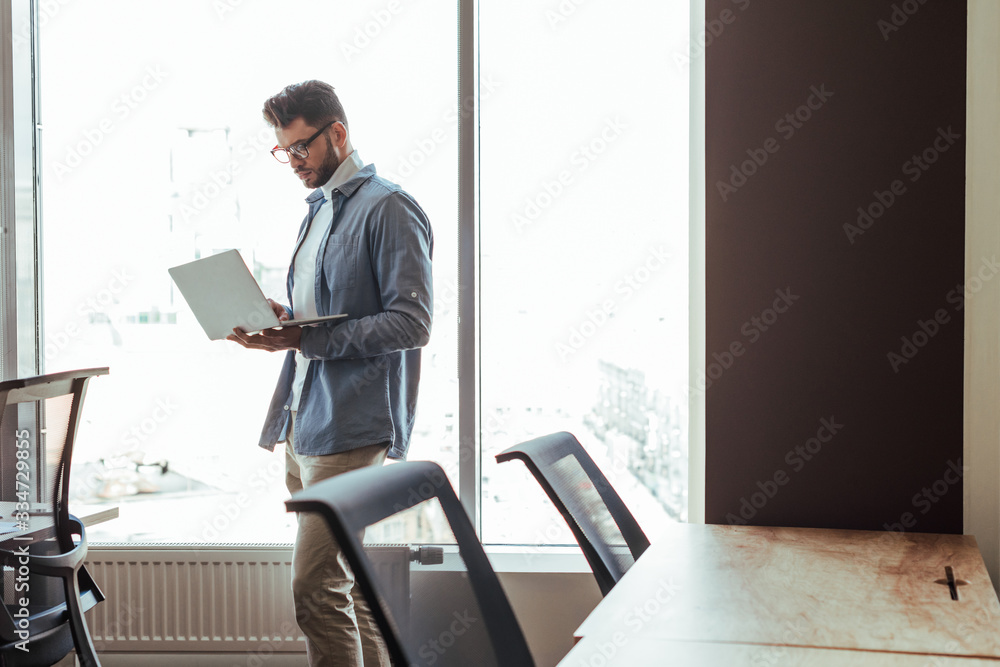 IT worker with laptop near windows in coworking space IT worker with laptop near windows in coworking space