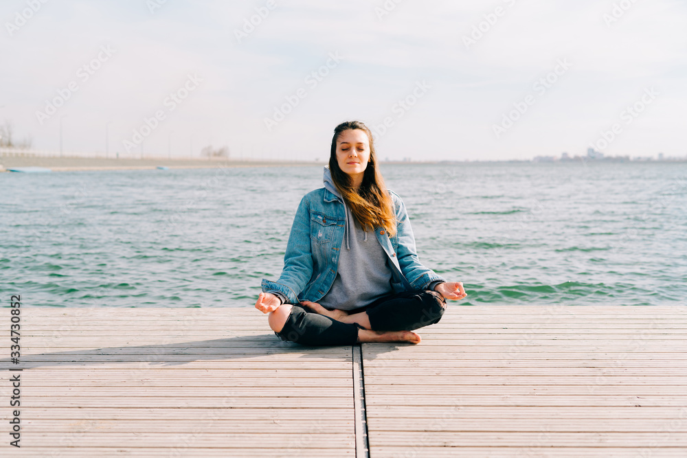 a young beautiful girl is sitting on a pier near the lake and doing meditation practice on a Sunny spring day