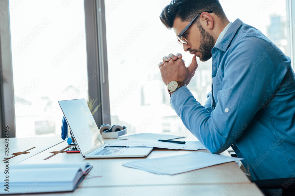 Concentrated IT worker with clenched hands looking at laptop at table in coworking space Concentrated IT worker with clenched hands looking at laptop at table in coworking space