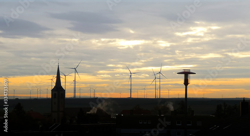 Scenic cloudy sky scape of dawn or sunset with dark silhouette of small German town on bottom line of image with tower of Catholic chapel,street lamp,wind turbines on horizon line, in contour light