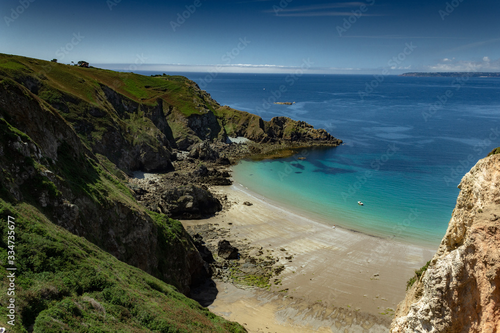 Coast and beach during sunny day with blue and turquoise waters surrounded by green and rocky cliffs in Sark, Guernsey, Channel Islands, United Kingdom