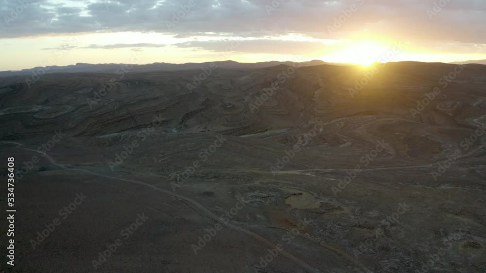 An slow aerial push on a narrow path winding through a stark, barren desert landscape dotted with interesting colours and geological features, at sunrise or sunset. An slow aerial push on a narrow path winding through a stark, barren desert landscape dotted with interesting colours and geological features, at sunrise or sunset.