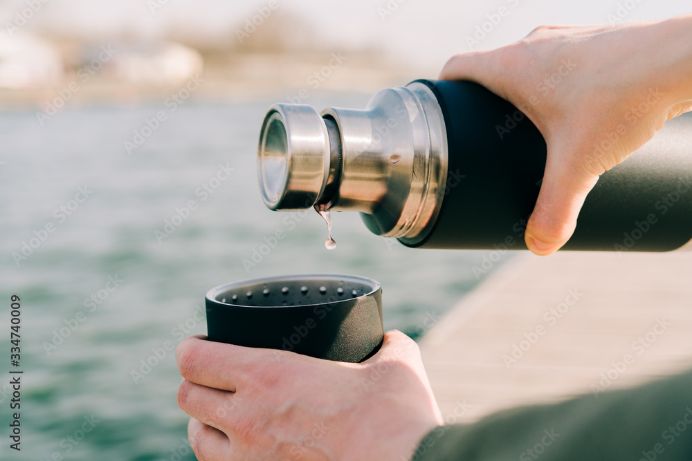 the guy is holding a black stylish eco-friendly stainless steel thermos and pours hot, herbal tea into a mug outdoors on a Sunny spring day near the lake the guy is holding a black stylish eco-friendly stainless steel thermos and pours hot, herbal tea into a mug outdoors on a Sunny spring day near the lake