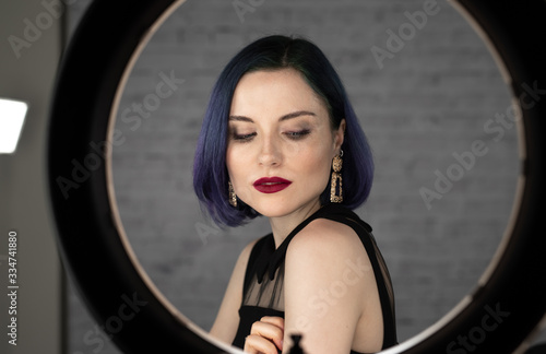 Young beautiful girl with professional makeup, blue hair and perfect skin sits in studio in front of ring light lamp posing for social media. Beauty blog. Copy space.