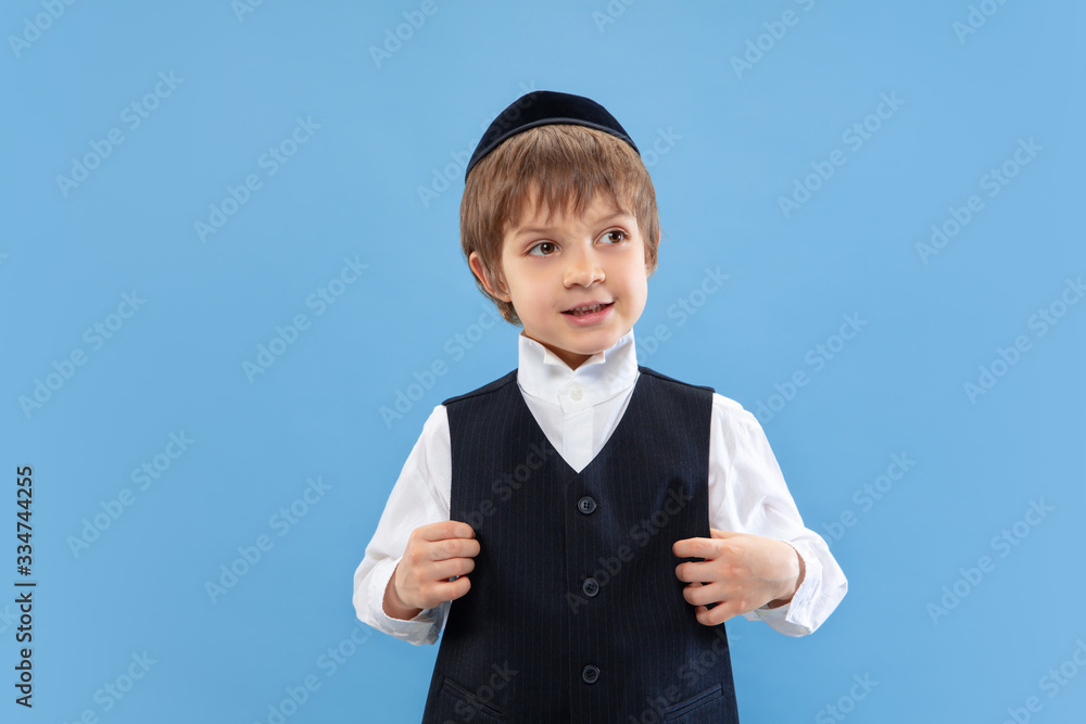 Posing confident, cute. Portrait of a young orthodox jewish boy isolated on blue studio background. Purim, business, festival, holiday, celebration Pesach or Passover, judaism, religion concept. Posing confident, cute. Portrait of a young orthodox jewish boy isolated on blue studio background. Purim, business, festival, holiday, celebration Pesach or Passover, judaism, religion concept.