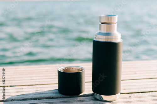 a beautiful black thermos of hot, herbal tea made from eco- friendly stainless steel stands on a wooden pier near the lake on a Sunny day