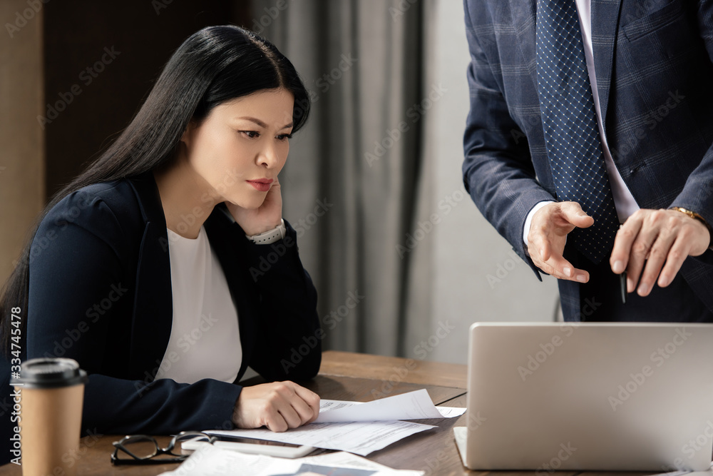 cropped view of businessman pointing with finger at laptop and asian businesswoman looking at it during business meeting cropped view of businessman pointing with finger at laptop and asian businesswoman looking at it during business meeting
