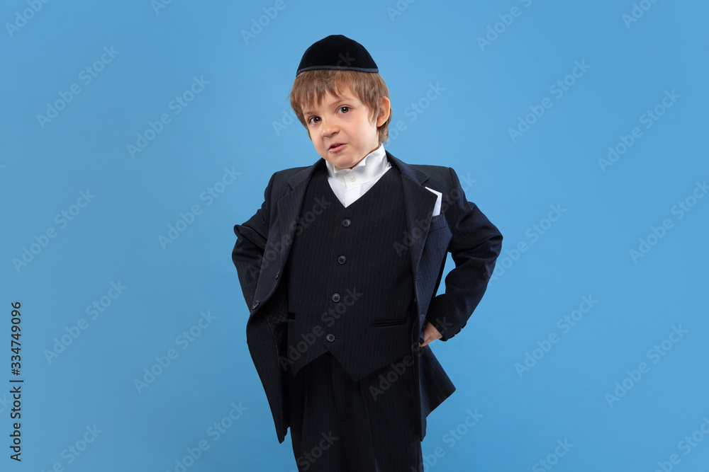 Posing confident, cute. Portrait of a young orthodox jewish boy isolated on blue studio background. Purim, business, festival, holiday, celebration Pesach or Passover, judaism, religion concept. Posing confident, cute. Portrait of a young orthodox jewish boy isolated on blue studio background. Purim, business, festival, holiday, celebration Pesach or Passover, judaism, religion concept.