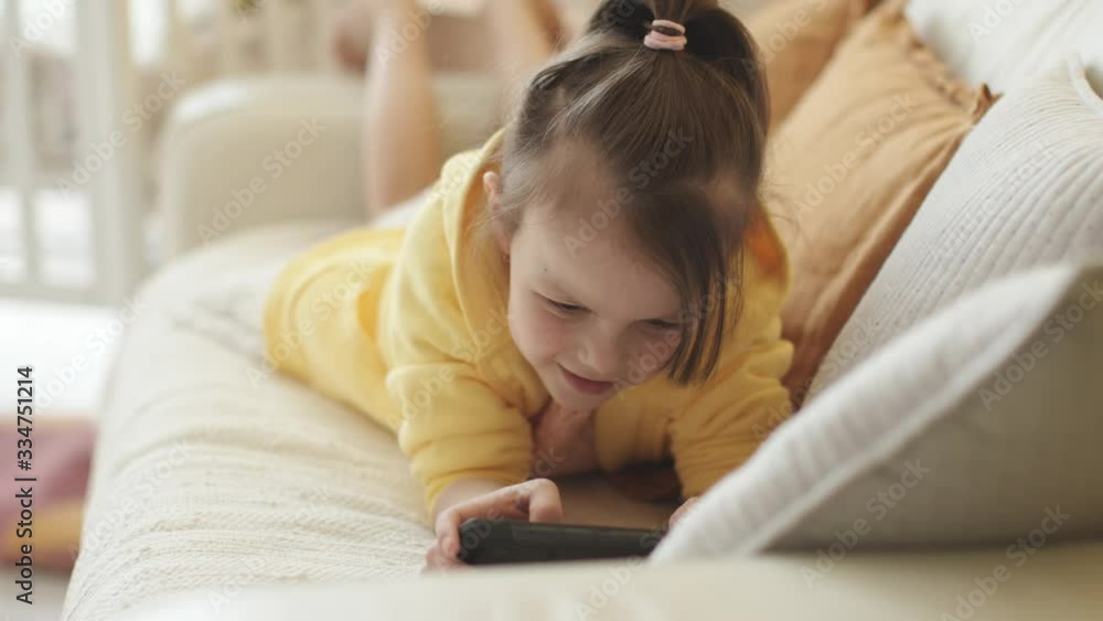 Little Girl in Yellow Hoodie Lying on Sofa and Holding Smartphone in her Hands, Playing Mobile Video Game. Sunny Yellow Cozy Home Interior. Little Girl in Yellow Hoodie Lying on Sofa and Holding Smartphone in her Hands, Playing Mobile Video Game. Sunny Yellow Cozy Home Interior.