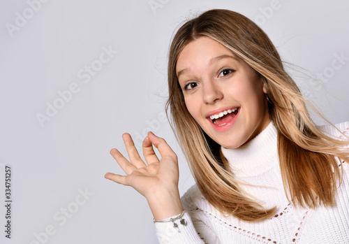 Blonde teenage female in sweater and bracelet. She is smiling and showing okay sign, posing isolated on white. Close up