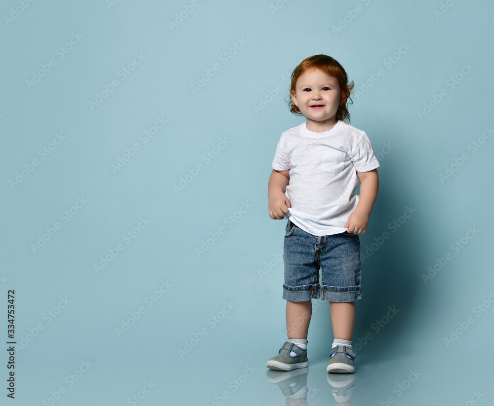 Little ginger toddler male or female in white t-shirt, socks and shoes,  denim shorts. Child is smiling, posing on blue background Stock Photo |  Adobe Stock