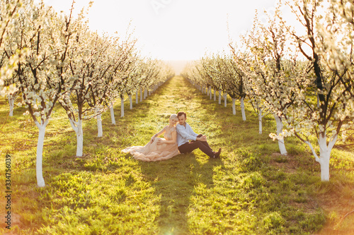 Happy smiling wedding couple in beautiful blooming garden, enjoying spring nature, sitting back to back on the green grass between rows of flowering trees. Love in the air, spring wedding concept © sofiko14