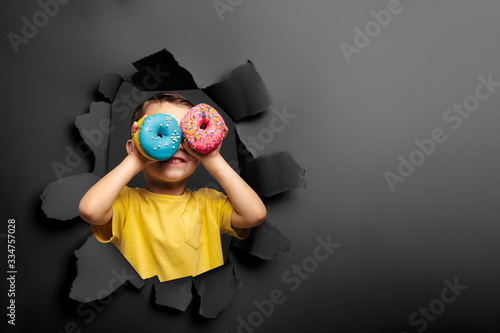 Happy cute boy is having fun played with donuts on black background wall.