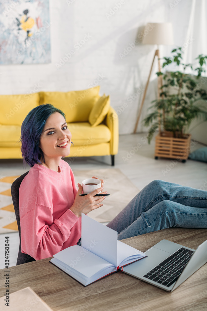 High angle view of freelancer with colorful hair, cup of tea and pen smiling and putting legs on table in living room High angle view of freelancer with colorful hair, cup of tea and pen smiling and putting legs on table in living room