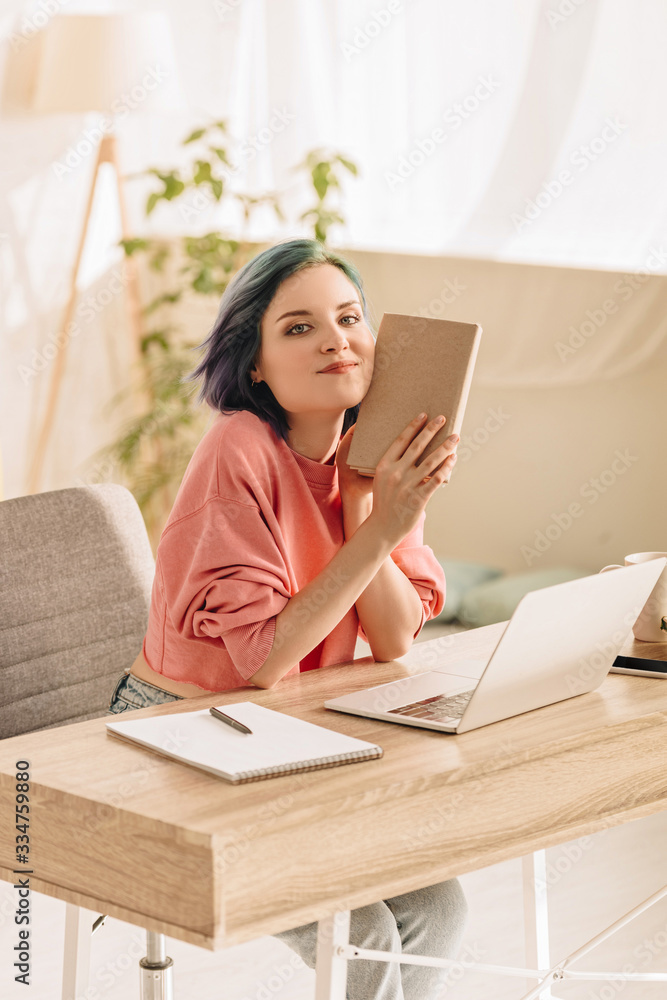 Freelancer with colorful hair holding book, looking at camera and smiling near laptop and notebook at table in living room Freelancer with colorful hair holding book, looking at camera and smiling near laptop and notebook at table in living room