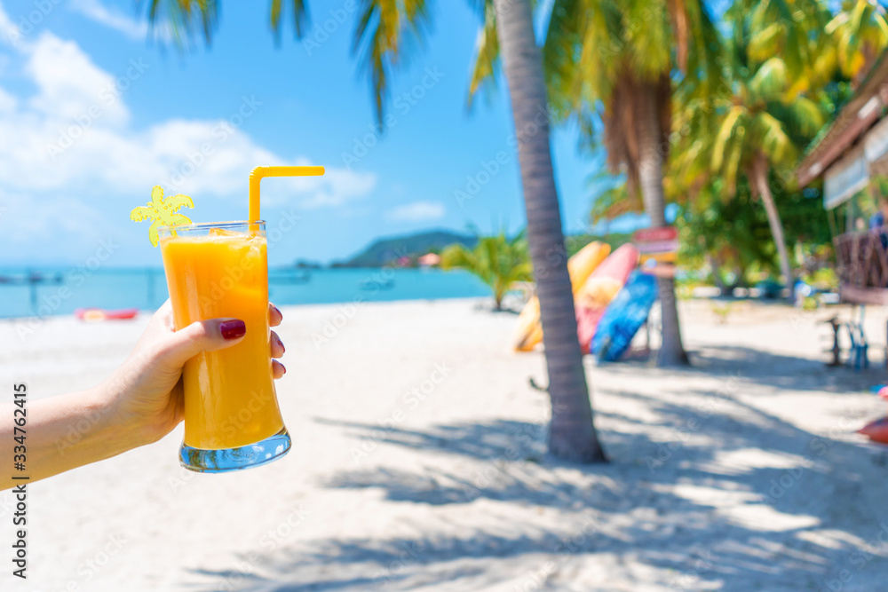 First-person view. Girl holds a glass cup of cold mango fresh on the background of a sandy tropical beach. White sand and palm trees. Fairytale vacation