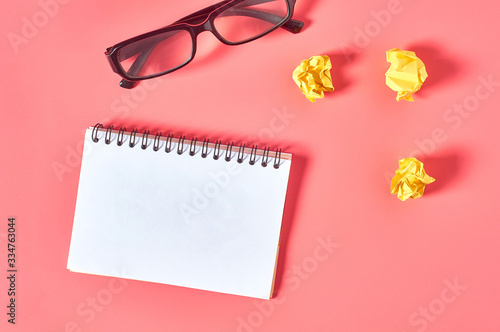Blank notepad near crumpled paper balls and modern glasses on pink desk. Education or business concept