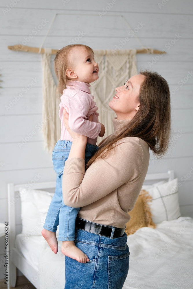 Mother and her daughter playing and laughing in their bedroom. Daughter in the arms of my mother. White bed linen, knitted blanket, macrame on the wall, hanging swing. Scandinavian style, cozy home. Mother and her daughter playing and laughing in their bedroom. Daughter in the arms of my mother. White bed linen, knitted blanket, macrame on the wall, hanging swing. Scandinavian style, cozy home.