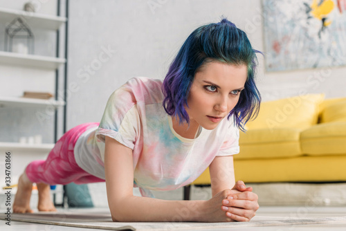 Selective focus of tense girl with colorful hair doing plank on yoga mat in living room
