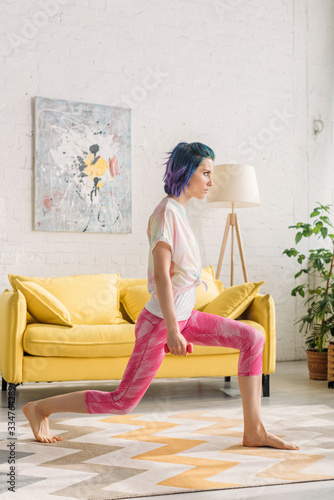 Beautiful woman with colorful hair doing lunges with dumbbell in living room