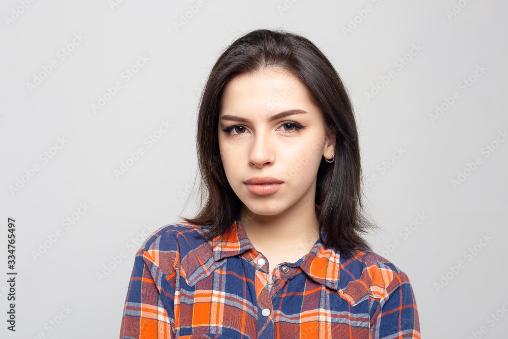 Portrait young beautiful woman not smiling in plaid shirt isolated on white-gray background. Girl with minimal natural make-up on her face. The photo is not retouched with natural defects on the skin. Portrait young beautiful woman not smiling in plaid shirt isolated on white-gray background. Girl with minimal natural make-up on her face. The photo is not retouched with natural defects on the skin.