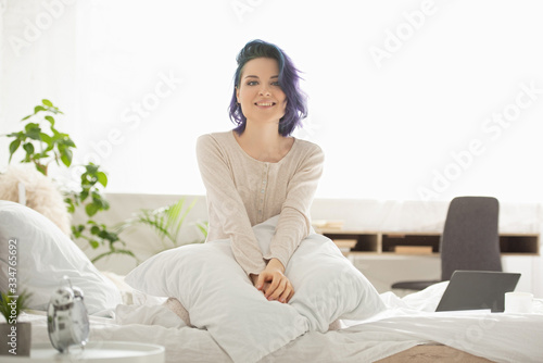 Beautiful woman with colorful hair looking at camera, smiling and sitting on bed in bedroom