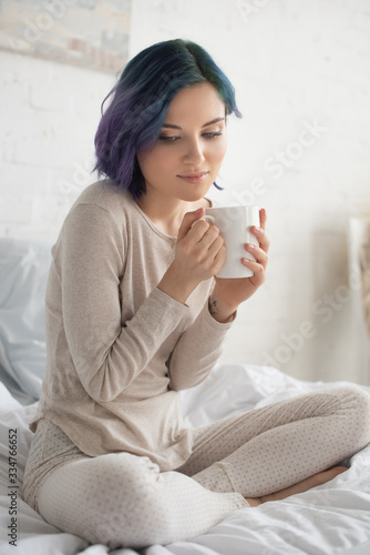 Thoughtful woman with colorful hair and crossed legs holding cup of tea on bed in bedroom