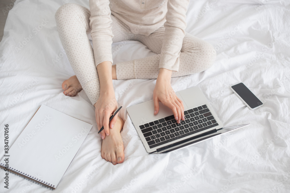 Partial view of freelancer with pen working on laptop near smartphone and notebook on bed Partial view of freelancer with pen working on laptop near smartphone and notebook on bed