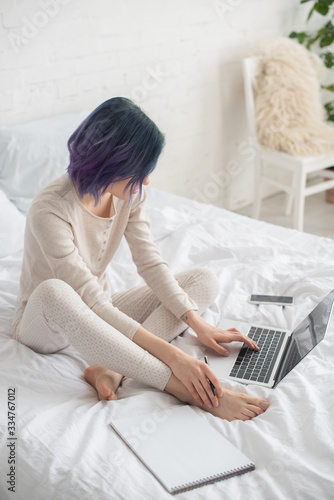 High angle view of freelancer with colorful hair holding pen and working on laptop near copybook on bed