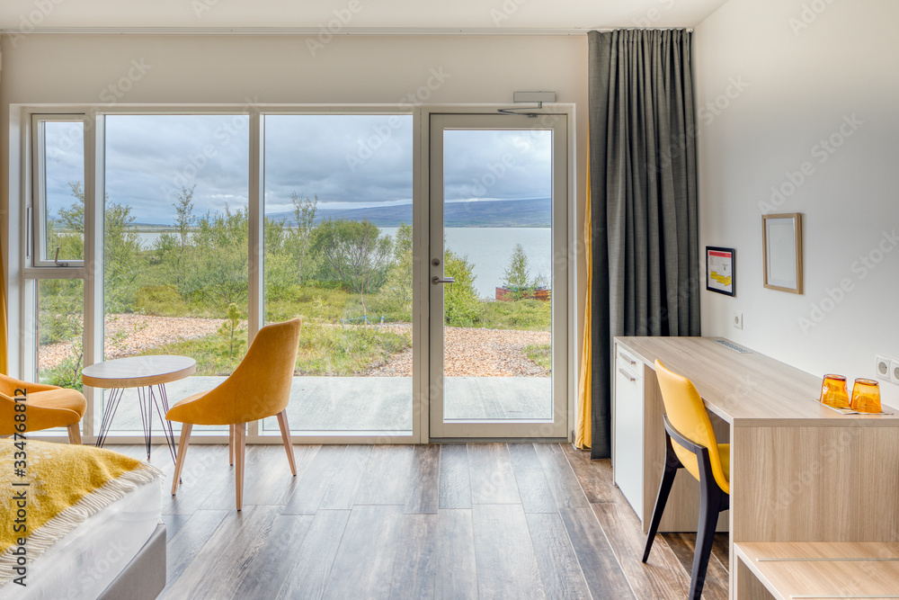 Outside views from inside a modern minimalist hotel room with chairs. Wooden tile floor. Plain white wall without decoration.. Huge windows. Closed glass door to the outside. Grey curtains Outside views from inside a modern minimalist hotel room with chairs. Wooden tile floor. Plain white wall without decoration.. Huge windows. Closed glass door to the outside. Grey curtains