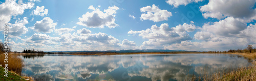 Panoramic landscape from the lake shore with colorful clouds in the spring sun. © mikhailgrytsiv