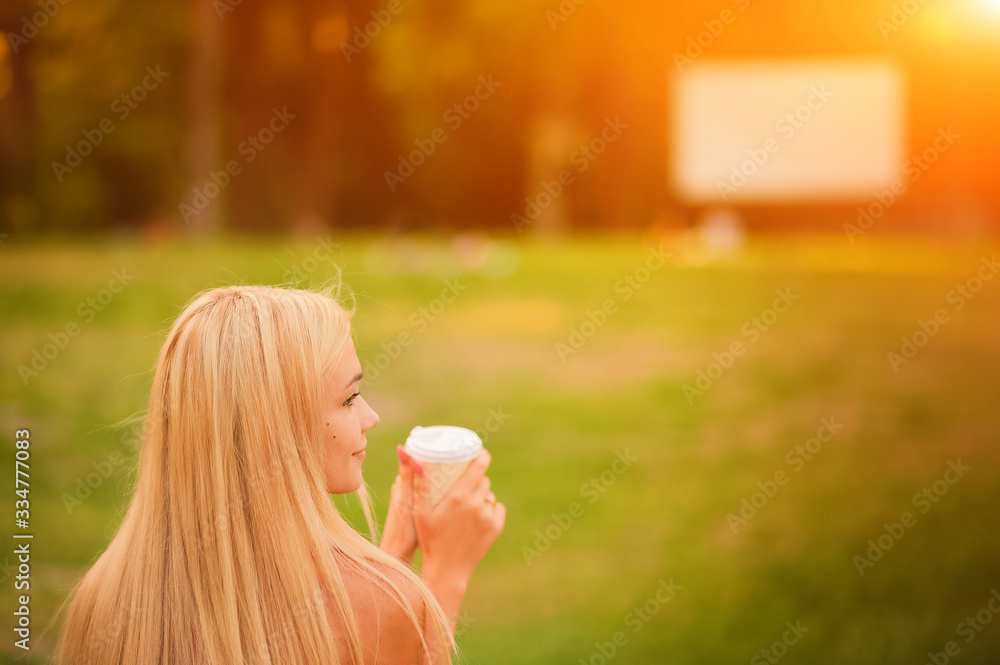 Girl on US Independence Day. A blonde is sitting on a lawn in front of an open-air cinema with coffee. USA Independence Day, America flag scarf