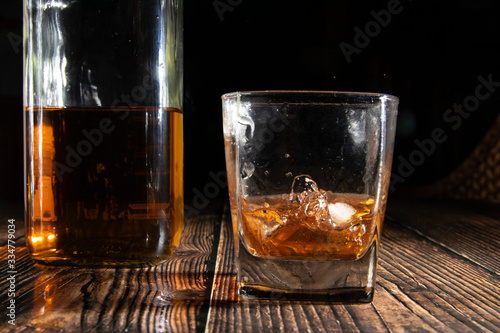 Glass of whiskey with ice cubes, a bottle on a wooden table. An old tabletop with light and a glass of strong drink.