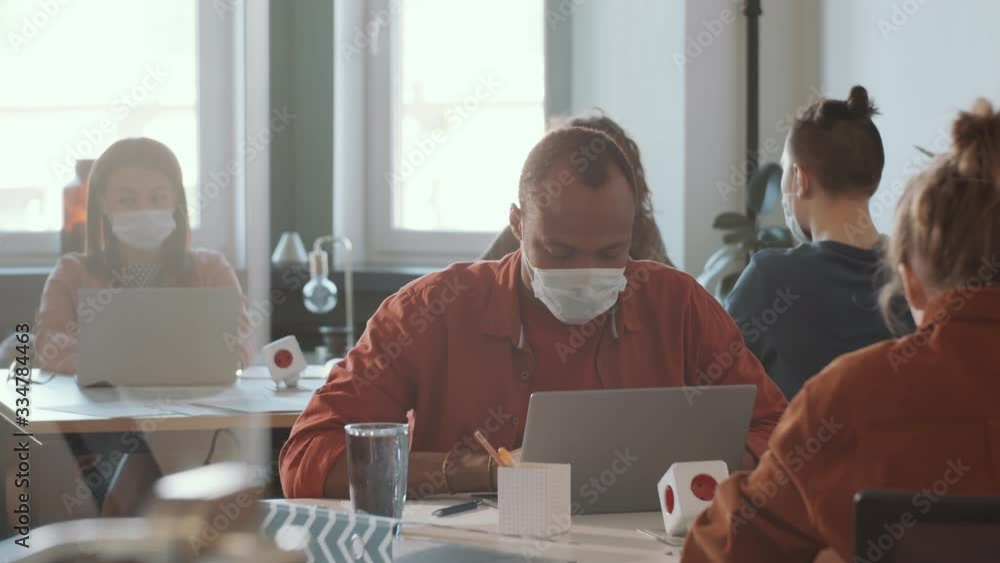 Young African American man in protective face mask sitting at table and using laptop while working in open space office with diverse colleagues during coronavirus outbreak Young African American man in protective face mask sitting at table and using laptop while working in open space office with diverse colleagues during coronavirus outbreak