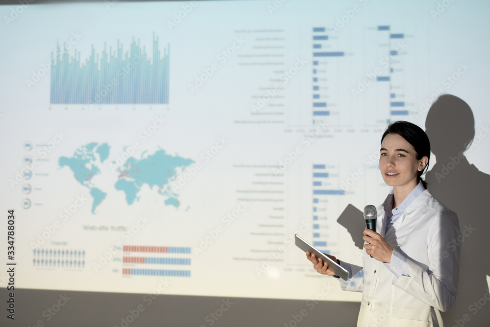 Content female medical student in lab coat using tablet while giving speech on coronavirus infection at conference Content female medical student in lab coat using tablet while giving speech on coronavirus infection at conference
