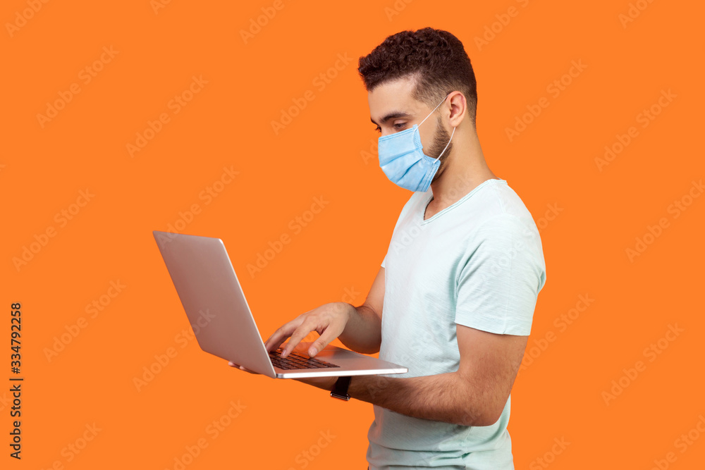 Side view portrait of cheerful brunette man with surgical medical mask working on laptop typing email or chatting in social network, happy face expression. studio shot isolated on orange background