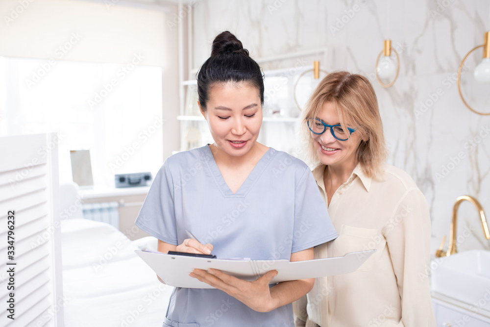 Asian beautician in scrubs filling information about client while talking to mature woman before cosmetic procedure Asian beautician in scrubs filling information about client while talking to mature woman before cosmetic procedure