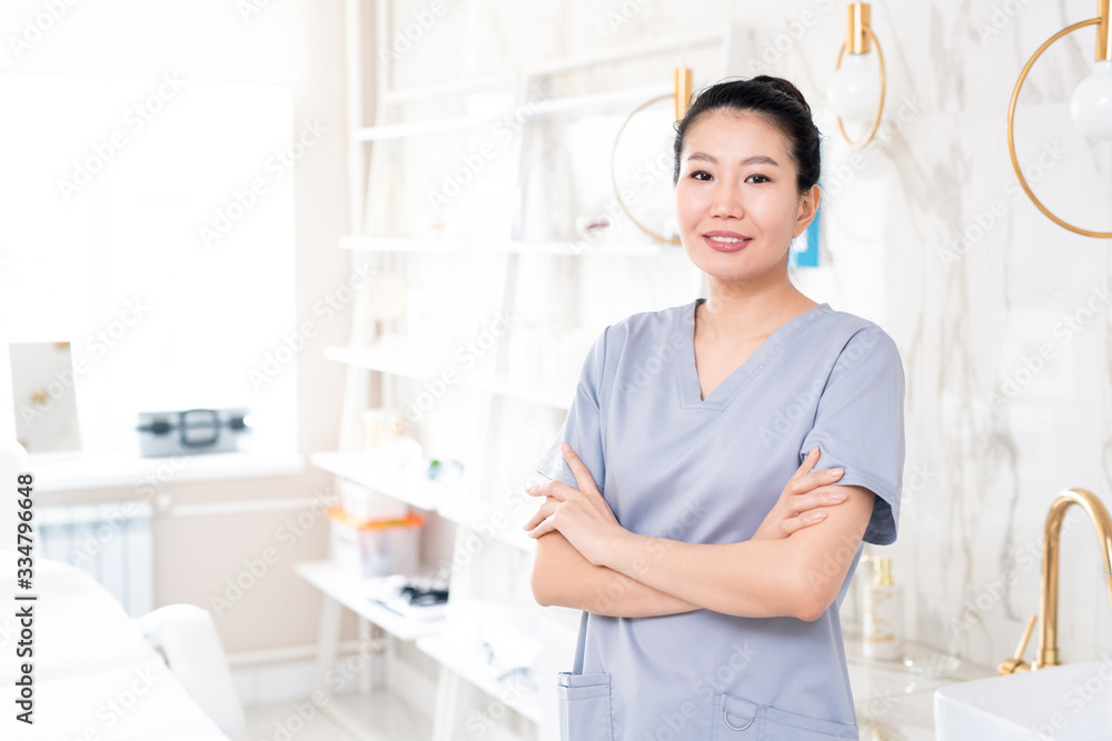 Portrait of positive middle-aged Asian beautician in scrubs standing with crossed arms in beauty treatment room with contemporary design Portrait of positive middle-aged Asian beautician in scrubs standing with crossed arms in beauty treatment room with contemporary design