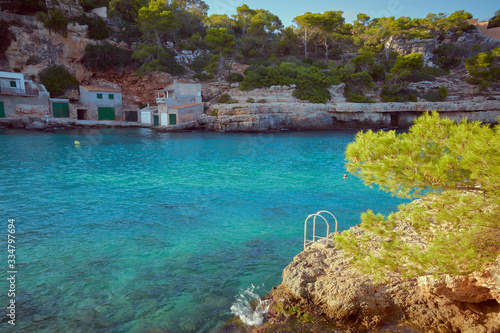Rocky beach in Mallorca. There are few folkloric houses in the background with many pines and in the foreground there is a metal staircase. Mallorca, Spain.