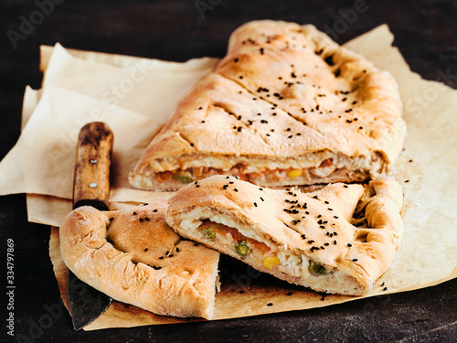 Perfect homemade closed pizza calzone on baking paper sheets over black background. Cutting tasty italian closed calzone pizza with cheese, meat, vegetables, black sesame dressed. Copy space for text