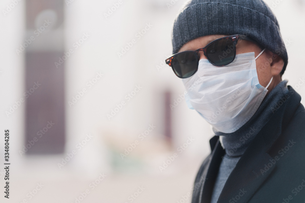 Coronavirus, quarantine and pandemic disease concept. Sideways shot of European man wears shades and disposable medical mask, poses outdoor against blurred background. Health care. Horizontal shot.