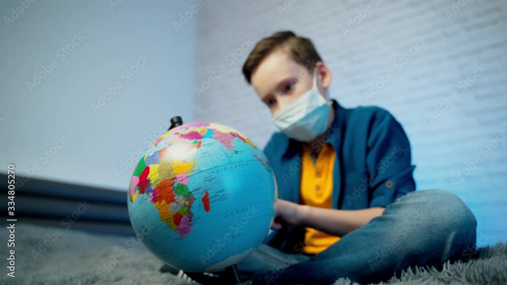 Boy looking at globe learning about virus spreading around the world. Boy wearing medical face mask. Concept of COVID-19 pandemic infection. Boy looking at globe learning about virus spreading around the world. Boy wearing medical face mask. Concept of COVID-19 pandemic infection.