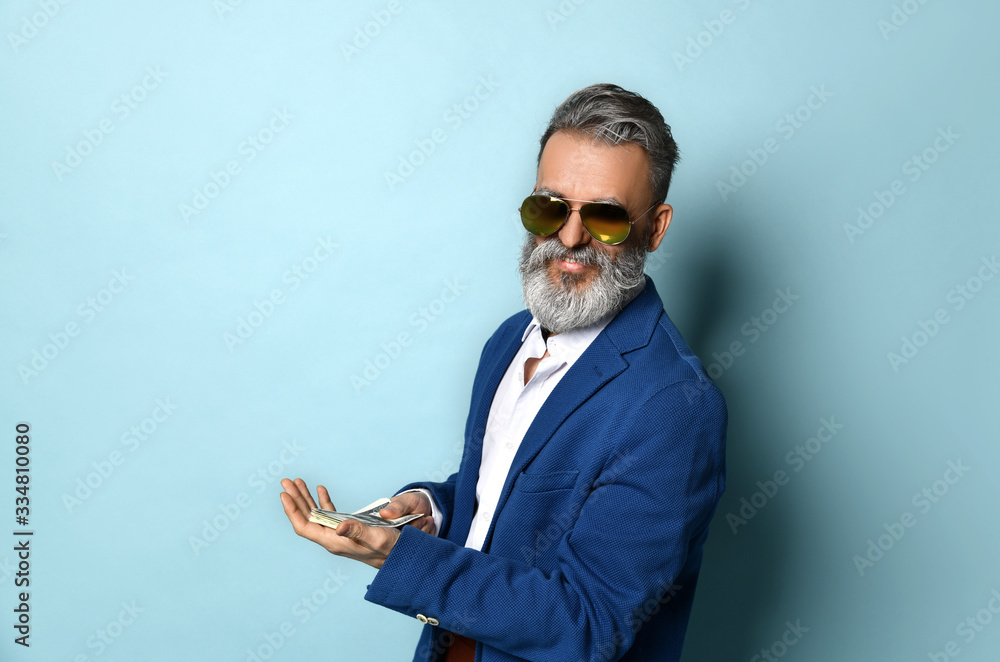 Elderly man in white shirt, jacket, brown pants and sunglasses. He showing fan of hundred dollar bills, posing on blue background
