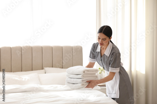 Young chambermaid putting stack of fresh towels in bedroom. Space for text photo