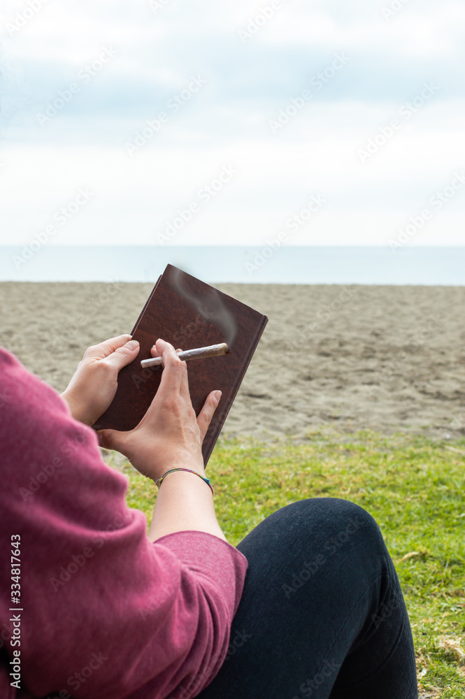 Person reading a book on beach take a break to smoke a marijuana joint or cigarette.