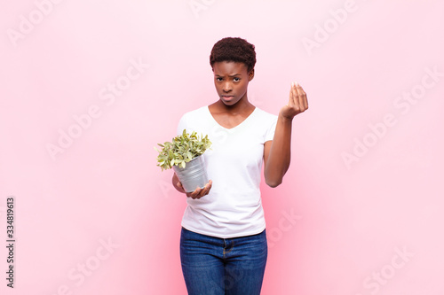 young pretty black womanmaking capice or money gesture, telling you to pay your debts! holding a plant photo