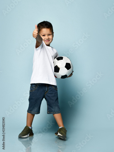 Little male in white t-shirt, shorts and khaki sneakers. Smiling, showing thumb up, holding soccer ball, posing on blue background