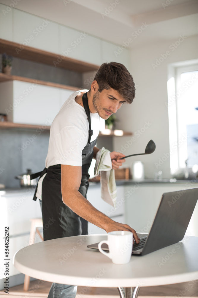 young man looking at laptop screen while cooking dinner young man looking at laptop screen while cooking dinner