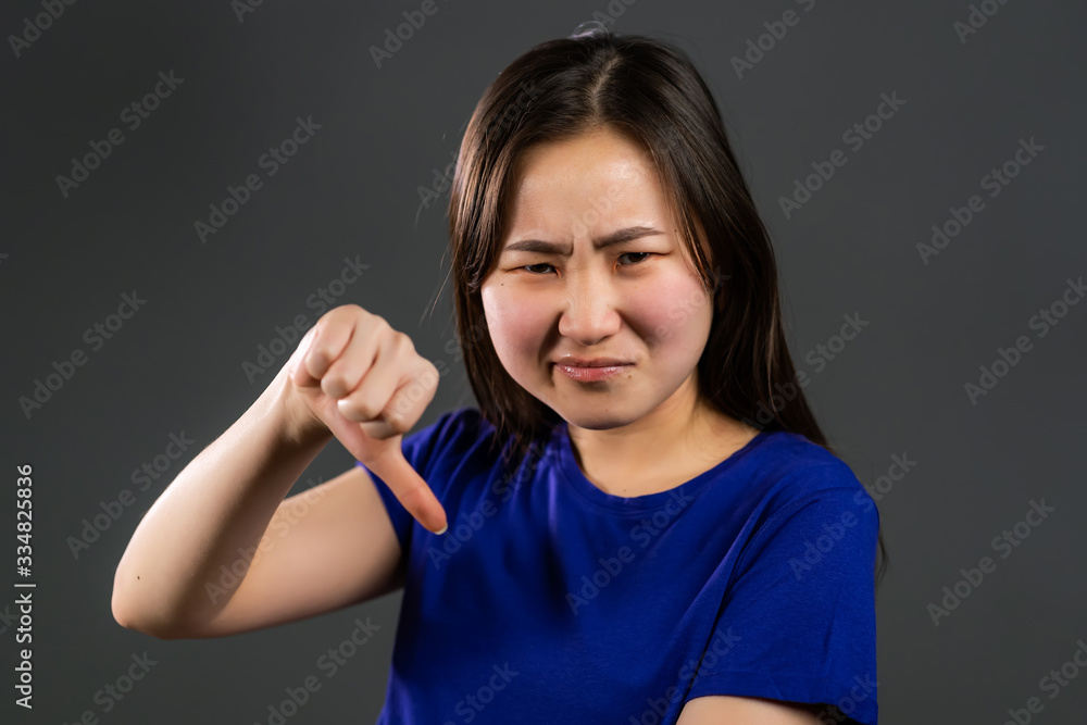 Fototapeta premium Young korean girl standing on grey studio background expressing discontent and showing thumb down gesture at camera. Portrait of asian woman with sign of dislike.