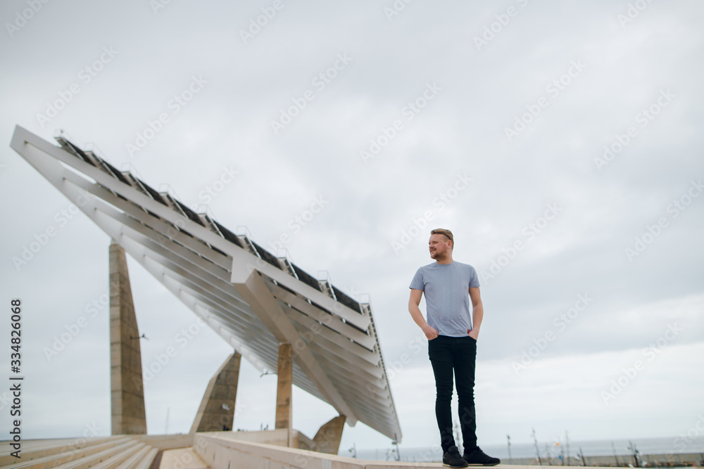 A young man in a t-shirt stands near big solar panel at Barcelona, Spain. Copy space